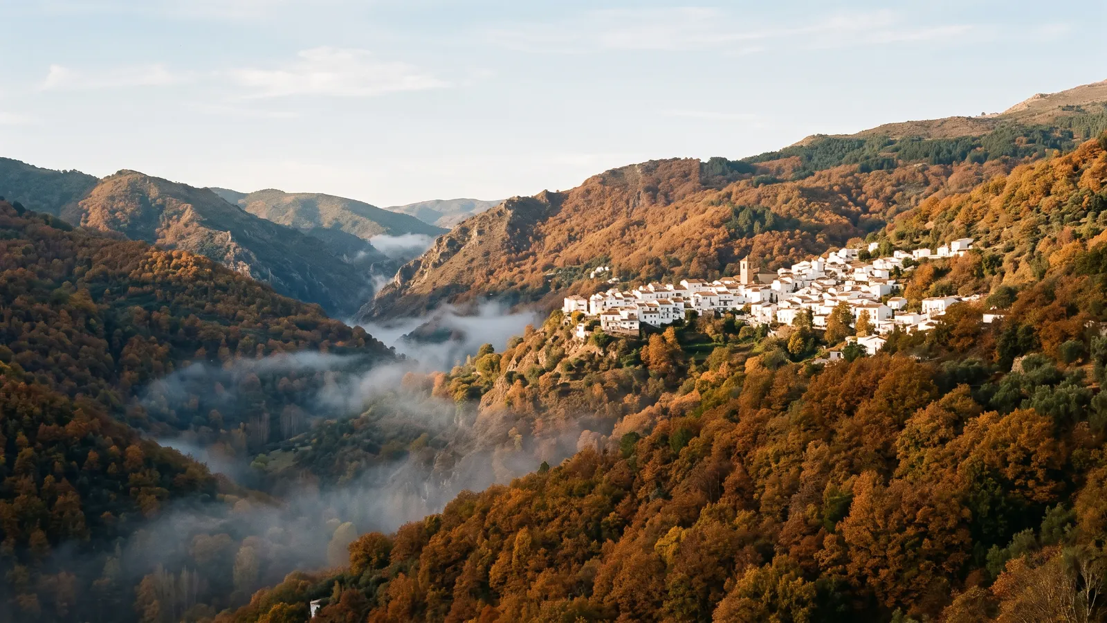 Valle de montaña en otoño con bosques de castaños cobrizos y un pueblo blanco en la ladera